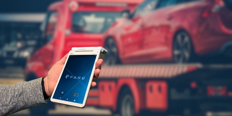 A hand holds a Fare terminal. There is a tow truck loaded up with a car in the background
